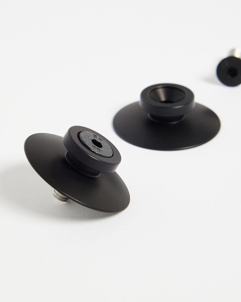 WoodUpp black anodized mounting discs with central threaded studs rest on a white background — two low-profile, matte flanged bases featuring raised collars and hex-key recessed caps, showing the screw thread underside; photographed close-up to highlight form, finish, and hardware details.