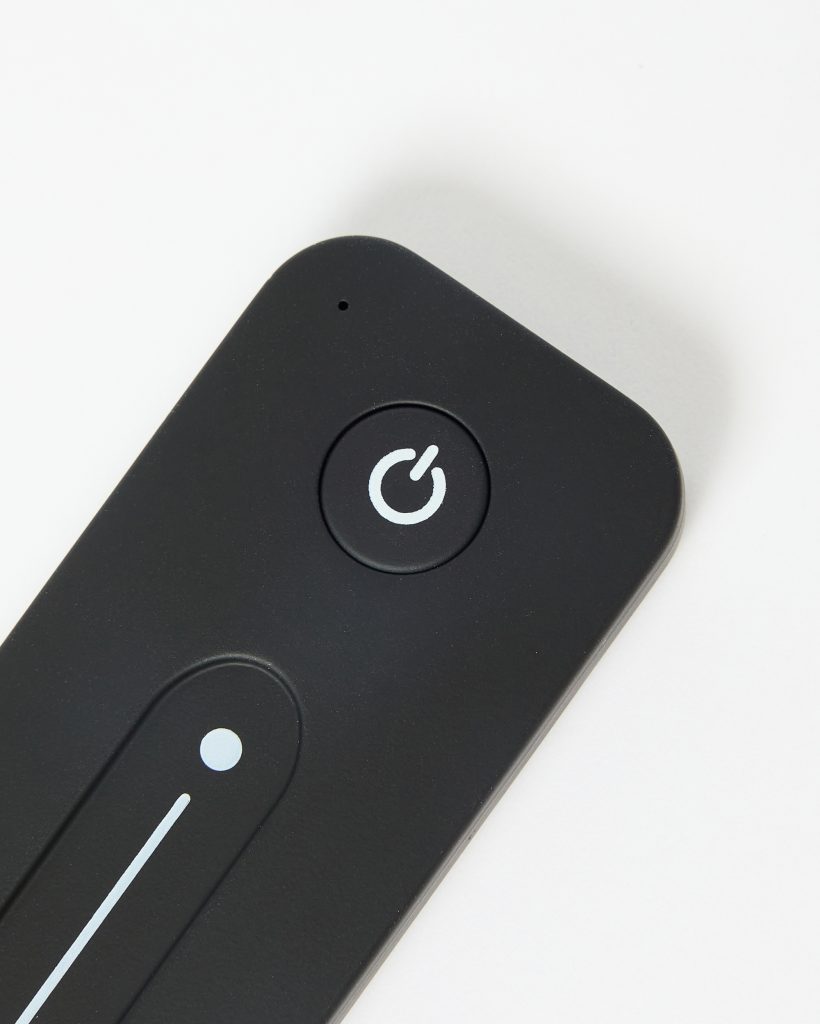 Close-up of a WoodUpp black remote control for LED lamps: matte, rounded rectangle featuring a circular power button with white power icon, an elongated touch dimmer strip with a small indicator dot and thin white line, plus a tiny sensor hole, on a white background.