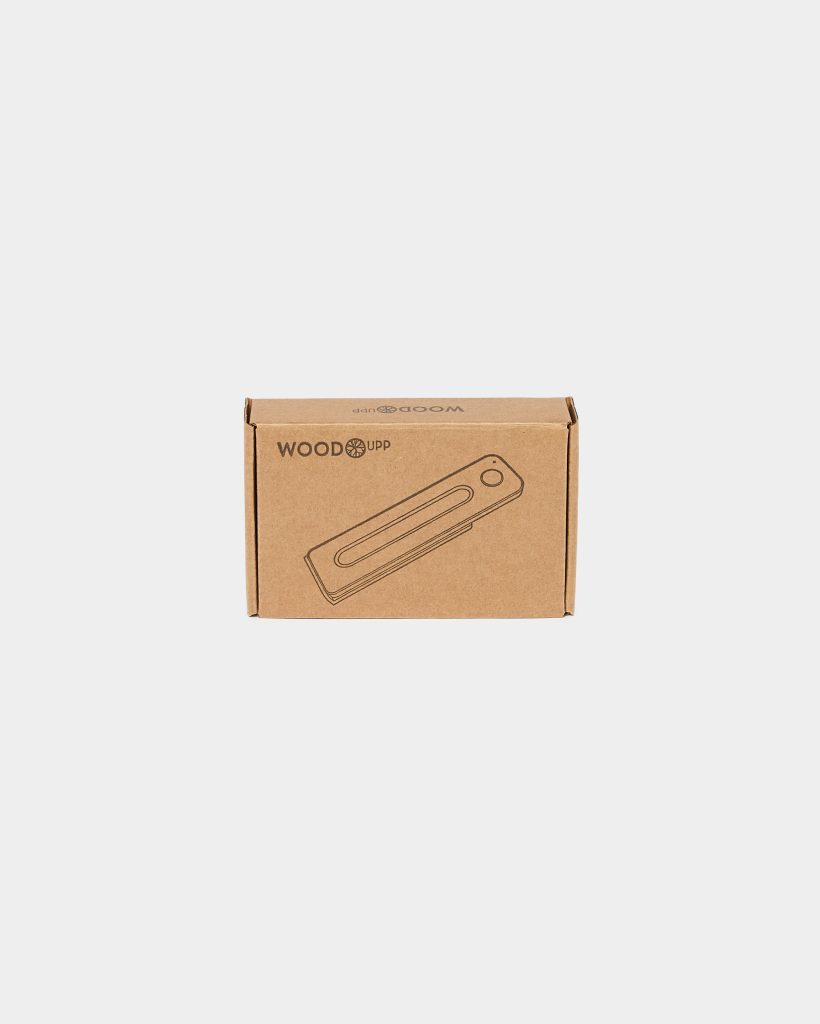 Centered on a white background, a WoodUpp brown cardboard box displays the WoodUpp logo and a printed line drawing of a slim wooden phone stand — a rectangular dock with a long central slot and a circular hole near one end, suggesting cable management.