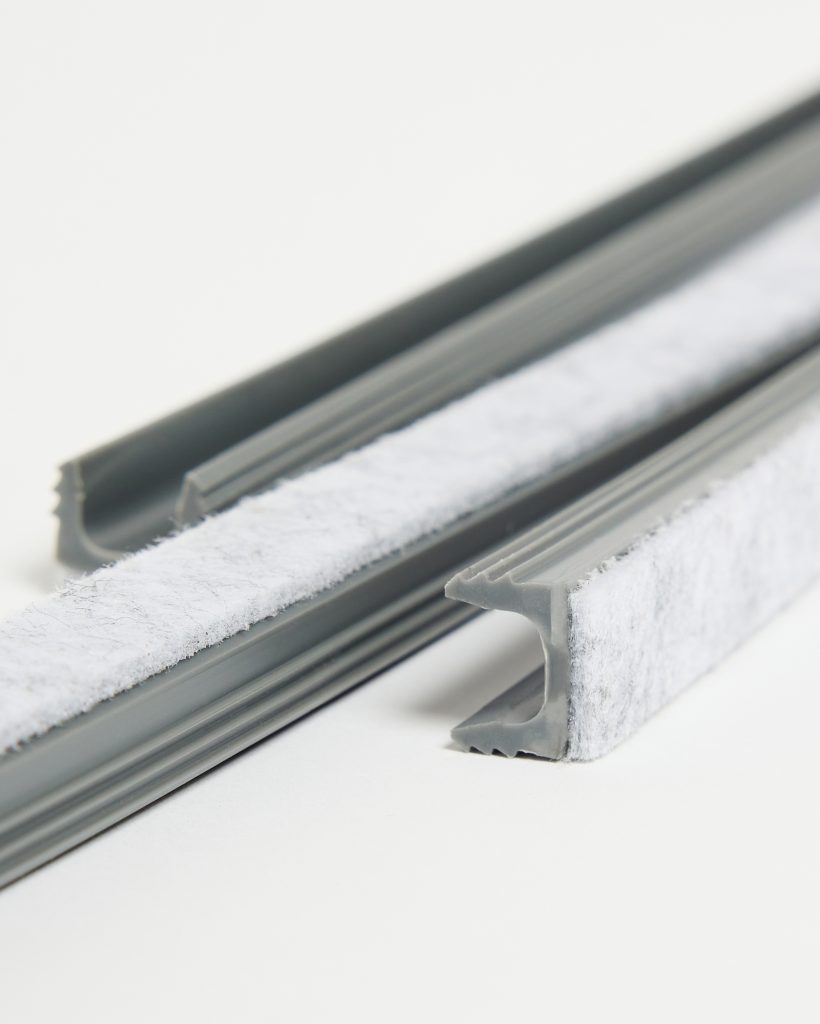 Close-up of WoodUpp grey pile weatherstrips: two extruded grey rubber channels fitted with soft white fibrous pile seals. The image shows U-shaped profiles, serrated gripping fins and the plush felt surface used for door and window drafts, demonstrating product texture and installation-ready ends.