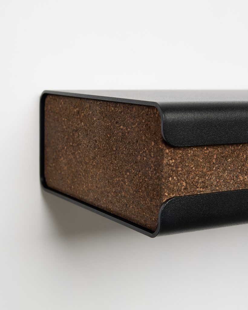 Close-up of WoodUpp wall shelf: a minimalist floating shelf featuring a matte black powder-coated steel frame that wraps around a thick natural cork block insert. The image highlights the textured brown cork surface and curved metal edges, showcasing WoodUpp’s modern, tactile shelf design.