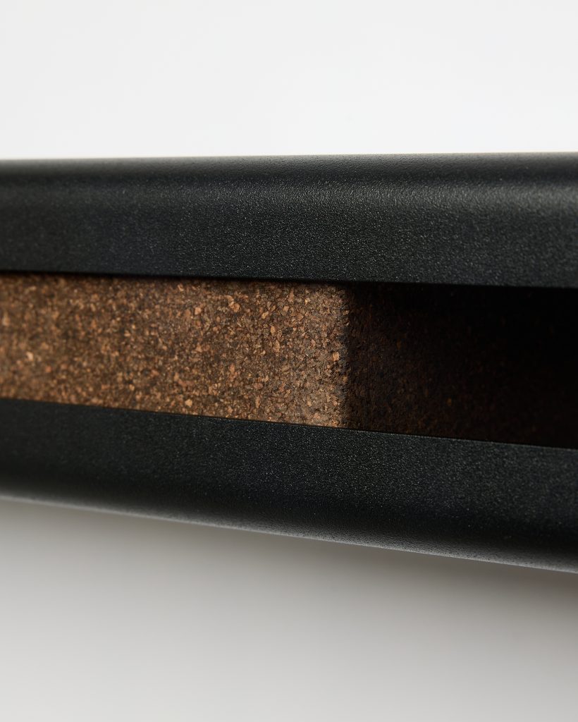 Close-up of a WoodUpp product: a matte black aluminium frame or rail holding a thick brown cork inlay. The image shows the textured cork block seated inside the sleek black metal channel, highlighting contrasting materials and the precise fit typical of WoodUpp’s desk accessory components.