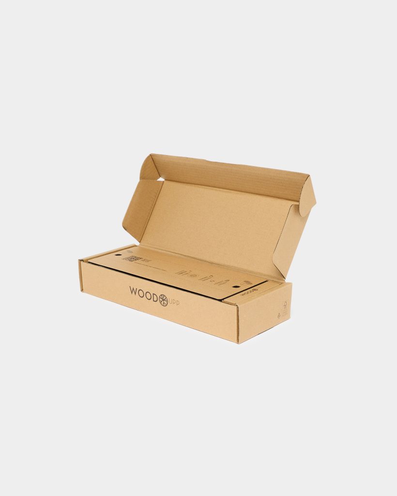 Open brown corrugated cardboard box branded WoodUpp, lid folded back to reveal a slim inner cardboard package printed with simple assembly icons and the WoodUpp logo; minimalist eco-friendly packaging photographed on white background, showing recyclable symbols and neat die-cut flaps.