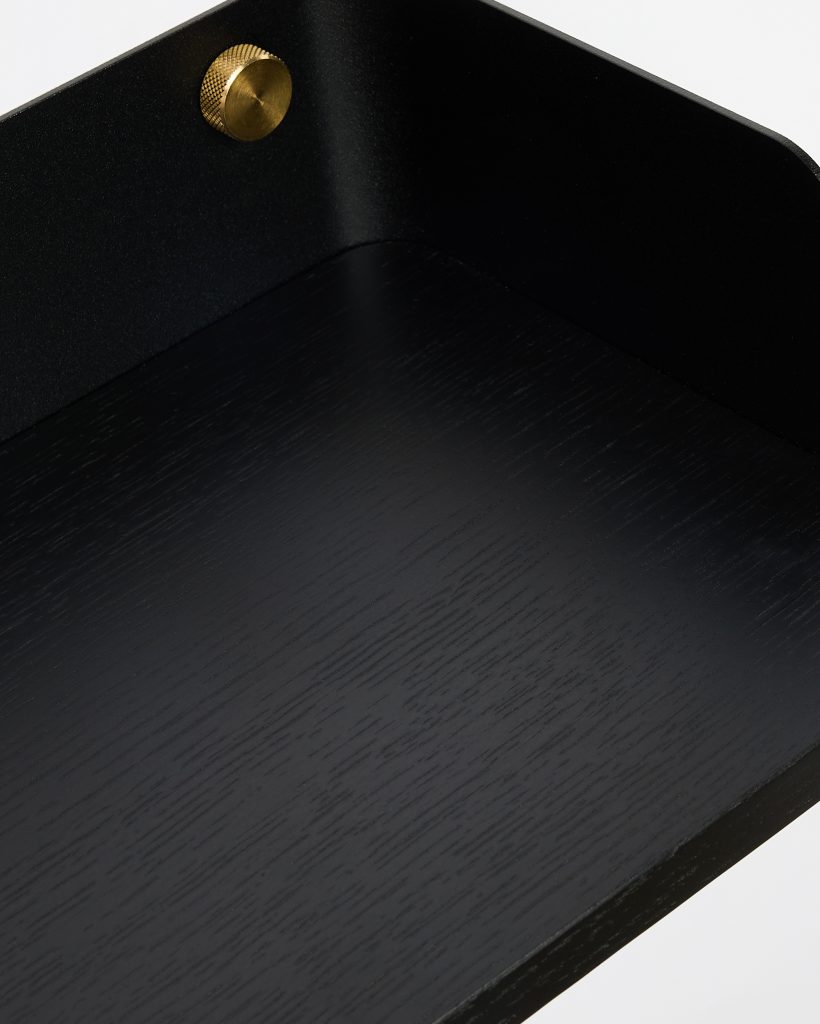 Close-up of a WoodUpp black oak shelf showing the wood grain on the flat surface, a matte black metal backplate forming a raised rear edge, and a knurled brass mounting screw. The image highlights the shelf’s textured finish, minimalist bracket design, and small decorative hardware.