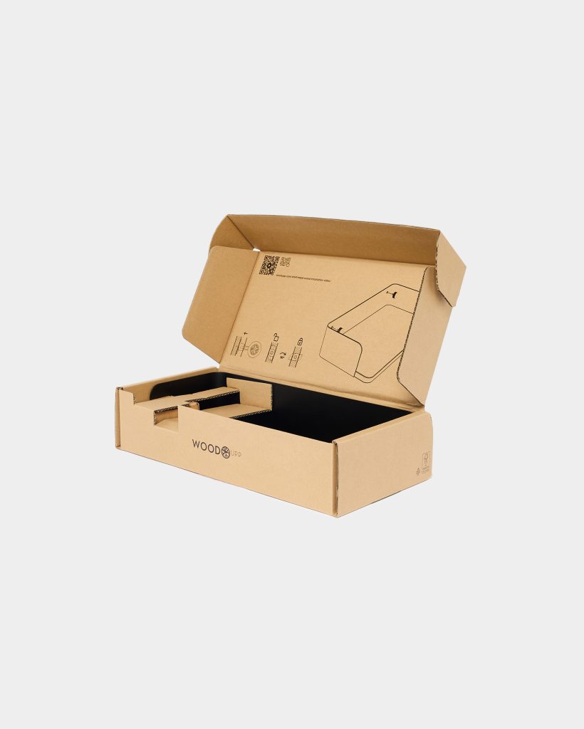 Open WoodUpp corrugated cardboard box with black internal insert and cardboard compartments, branded "WoodUpp" on the front. Inside lid shows a printed assembly diagram and QR code for a WoodUpp wooden desk stand, indicating packaging for a slim wooden laptop or monitor stand.