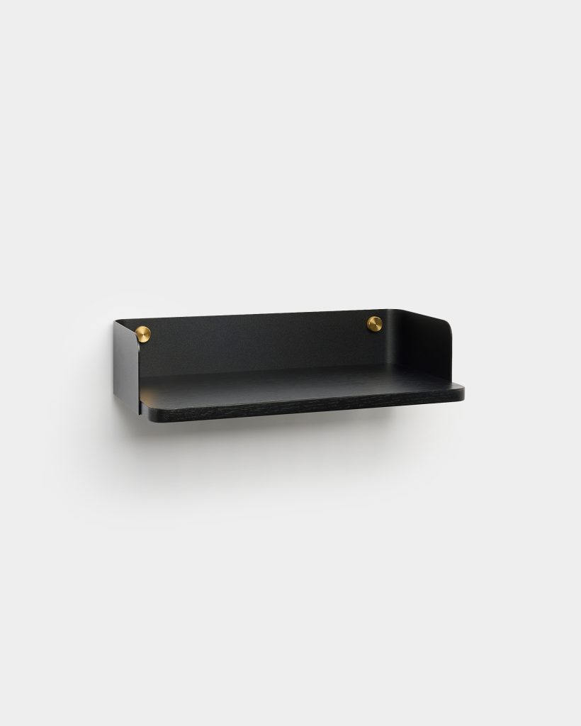 WoodUpp black wall shelf on white background, featuring a matte black metal rear panel with curved sides and a slim black wooden shelf, fastened by two round brass-finish studs. Minimal floating design ideal for small decor, keys or a phone.