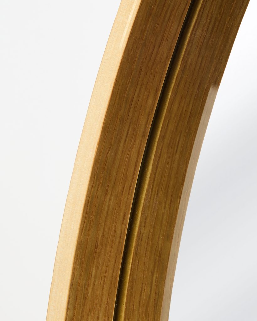 Close-up of a WoodUpp round mirror frame showing a curved layered plywood profile with warm oak veneer and visible fine wood grain, a contrasting pale inner ply, glossy lacquered surface, a slim brass-coloured inlay and the mirror’s reflective glass edge in a minimalist modern design.