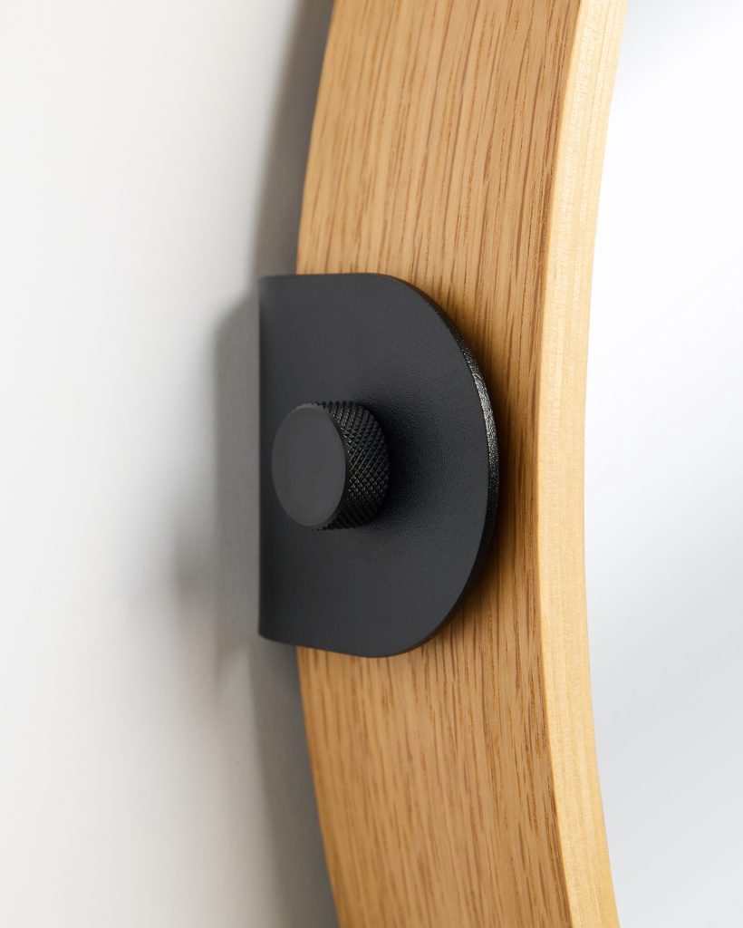 Close-up of a WoodUpp oak mirror frame showing its curved light-oak veneer grain and a matte black wall-mount bracket. The WoodUpp metal hanger features a rounded plate and knurled adjustment knob securing the frame to the wall, highlighting the contrasting wood texture and textured black finish.