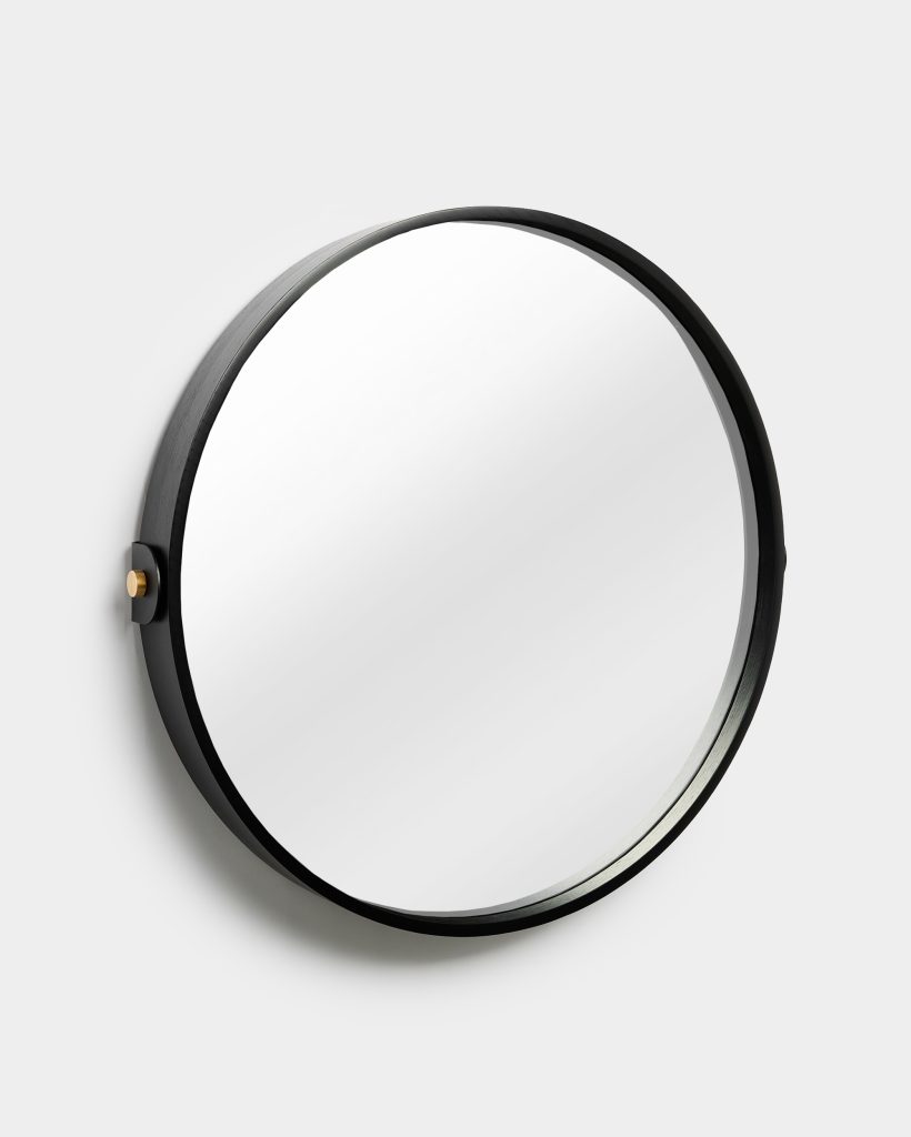 WoodUpp round wall mirror with thin black-stained wooden frame, circular glass face and a single visible brass mounting accent on the left; photographed against a neutral light-gray wall, showing the mirror's minimalist, contemporary design and slim profile, highlighting high-quality craftsmanship.