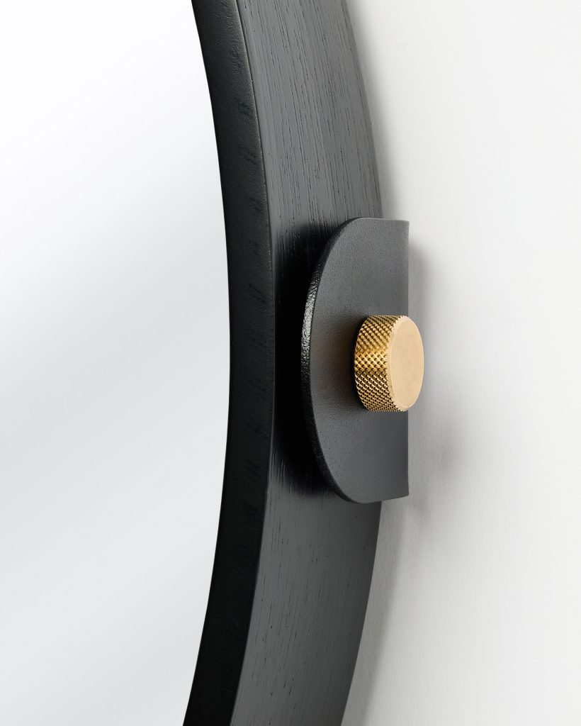 Close-up of a WoodUpp wall mirror showing the black wooden circular frame, matte black metal mounting bracket and distinctive knurled brass mounting knob attached to a white wall, highlighting the mirror’s wood grain, hardware texture and premium finish and craftsmanship.