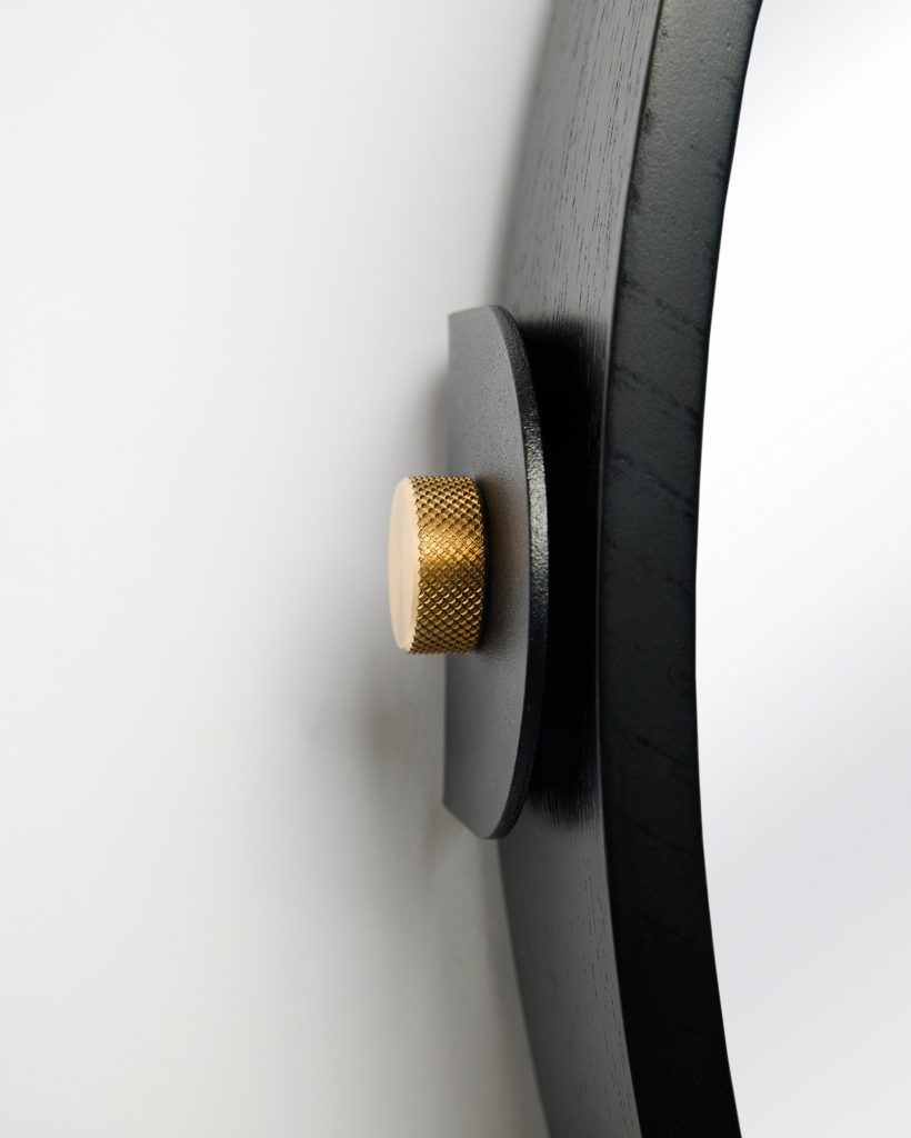 Close-up of a WoodUpp round mirror’s mounting: black-stained wooden frame and matte black metal wall bracket secured to the wall with a textured brass knurled screw knob. Visible are the mirror’s curved edge, subtle wood grain and minimalist hardware showcasing WoodUpp’s design details.