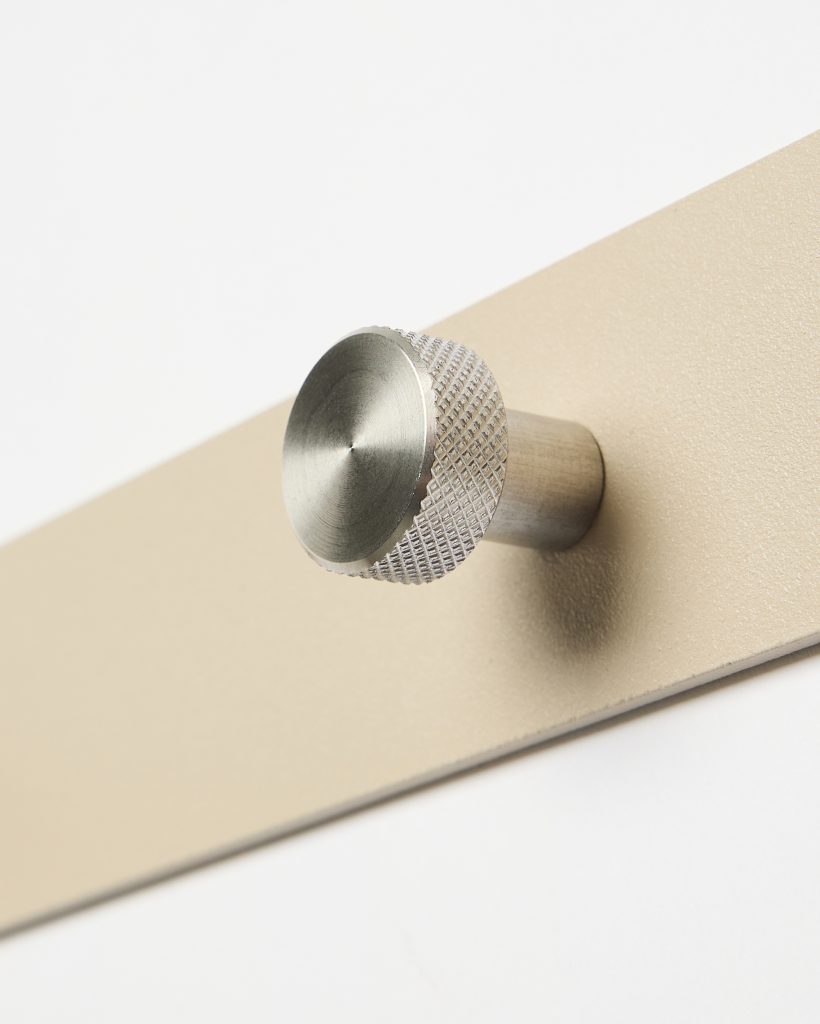WoodUpp metal wall hook: a single brushed stainless-steel peg with a circular, knurled-edge face and short cylindrical stem mounted on a matte sand-coloured rectangular backplate. Close-up shows textured machining on the knob and subtle brushed finish against the smooth powder-coated plate.