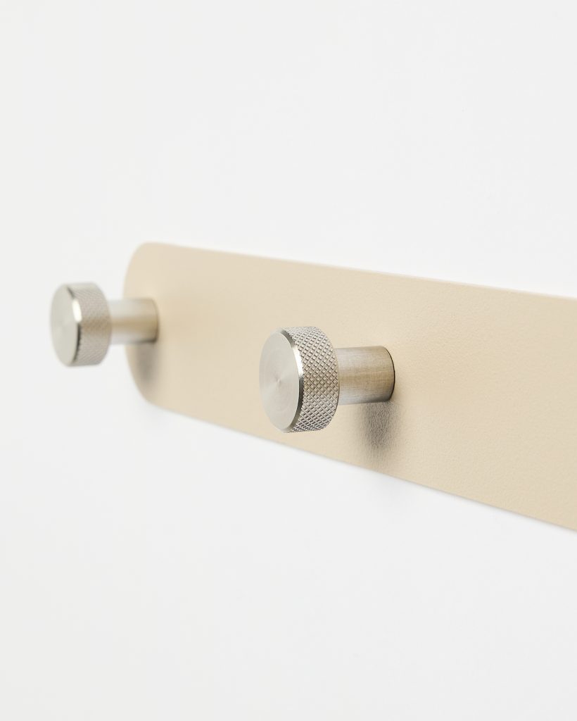 WoodUpp wall-mounted coat rack featuring a slim beige metal backplate with two brushed stainless steel cylindrical pegs. Each peg has a textured, knurled round head and short stem, shown in close-up against a white wall, highlighting minimalist Scandinavian design for hanging coats and accessories.