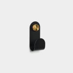 WoodUpp wall hook: a matte black, single metal coat hook with rounded top and sweeping curved loop, mounted by a knurled brass screw head. Minimal, sculptural design photographed on a light background, showing texture and contrasting brass fastener for storage.