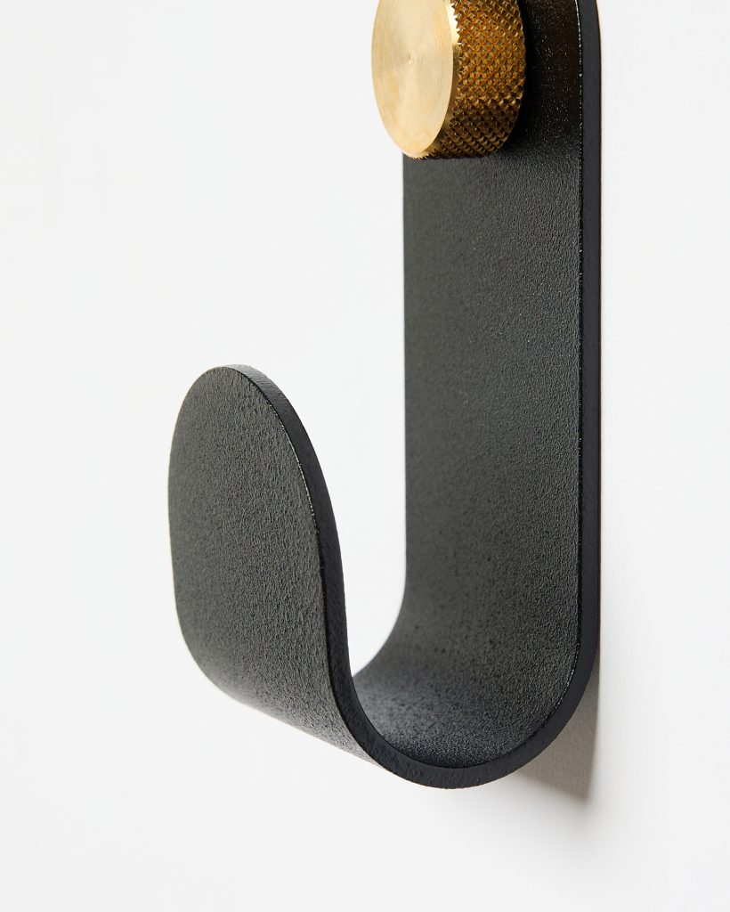 Close-up of a WoodUpp black matte curved wall hook with textured J-shaped profile, mounted on a white wall and fastened by a knurled brass WoodUpp mounting screw with circular head. Minimal, industrial design showing material texture and rounded hook profile.