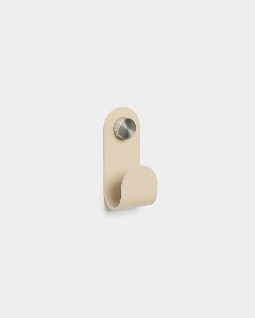 WoodUpp beige wall hook mounted on a white background, featuring a single compact curved metal hook with rounded top and smooth matte finish; secured by a knurled brushed stainless steel screw cap. Minimal, modern design suitable for coats, towels, bags, or keys.