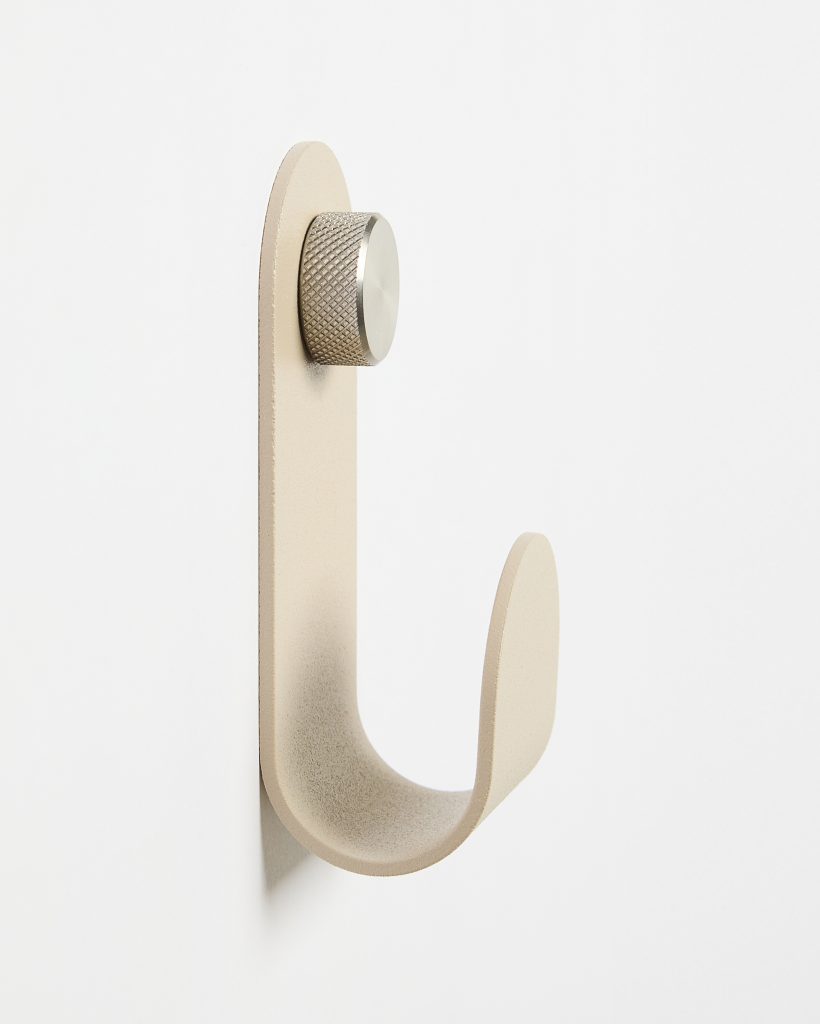 WoodUpp wall hook: a minimalist J-shaped hanger in matte sand beige with a smooth curved profile and subtle textured finish, secured to the wall by a knurled stainless-steel cap fastener. The compact, modern WoodUpp hook is suited for coats, bags or towels.