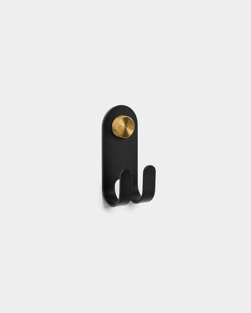 WoodUpp black metal double wall hook: a matte, rounded-rectangle backplate with two curved twin hooks and a knurled brass circular mounting knob. Minimalist, industrial-style design shown mounted on a plain white background, highlighting the compact, durable metal construction and decorative brass accent.
