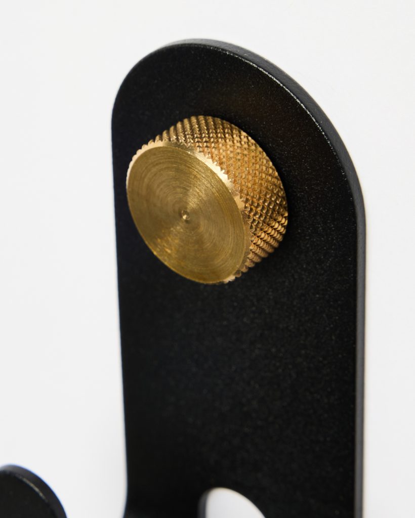 Close-up of a WoodUpp black matte metal wall hook showing a rounded top plate and a textured brass knurled screw-style peg; the solid circular brass knob serves as a decorative fastening and hanging peg against a white background, highlighting material contrast and precise machining.