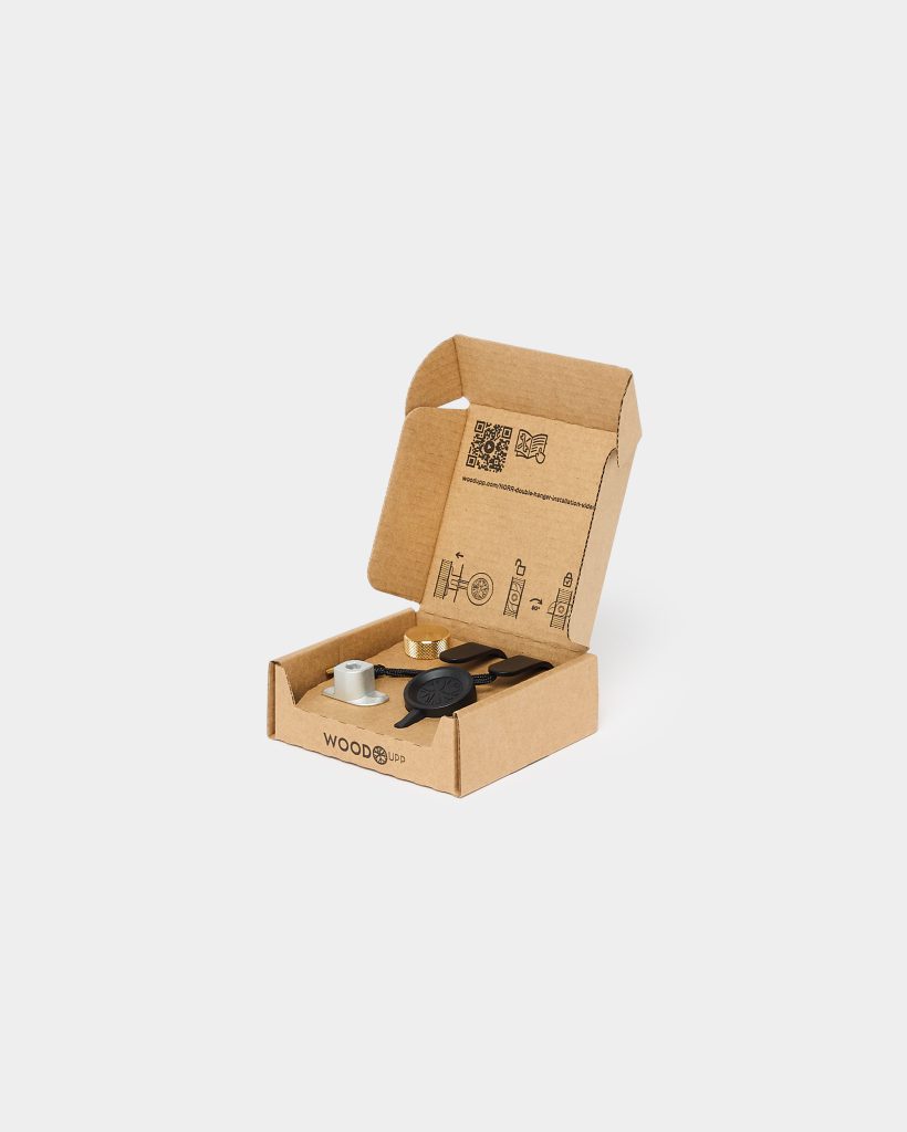 WoodUpp open cardboard box displaying a WoodUpp mounting kit: a brass knurled screw knob, a silver metal spacer, a black plastic wall mount with corded loop and two black clip anchors. Inside lid are printed assembly diagrams and a QR code on the recyclable packaging.
