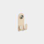 WoodUpp double wall hook mounted on a white background: a matte beige metal backplate with rounded top and two curved hooks, secured by a knurled brushed-steel fixing cap. Clean, minimalist design ideal for coats, bags or towels, showing product shape, finish and hardware detail.