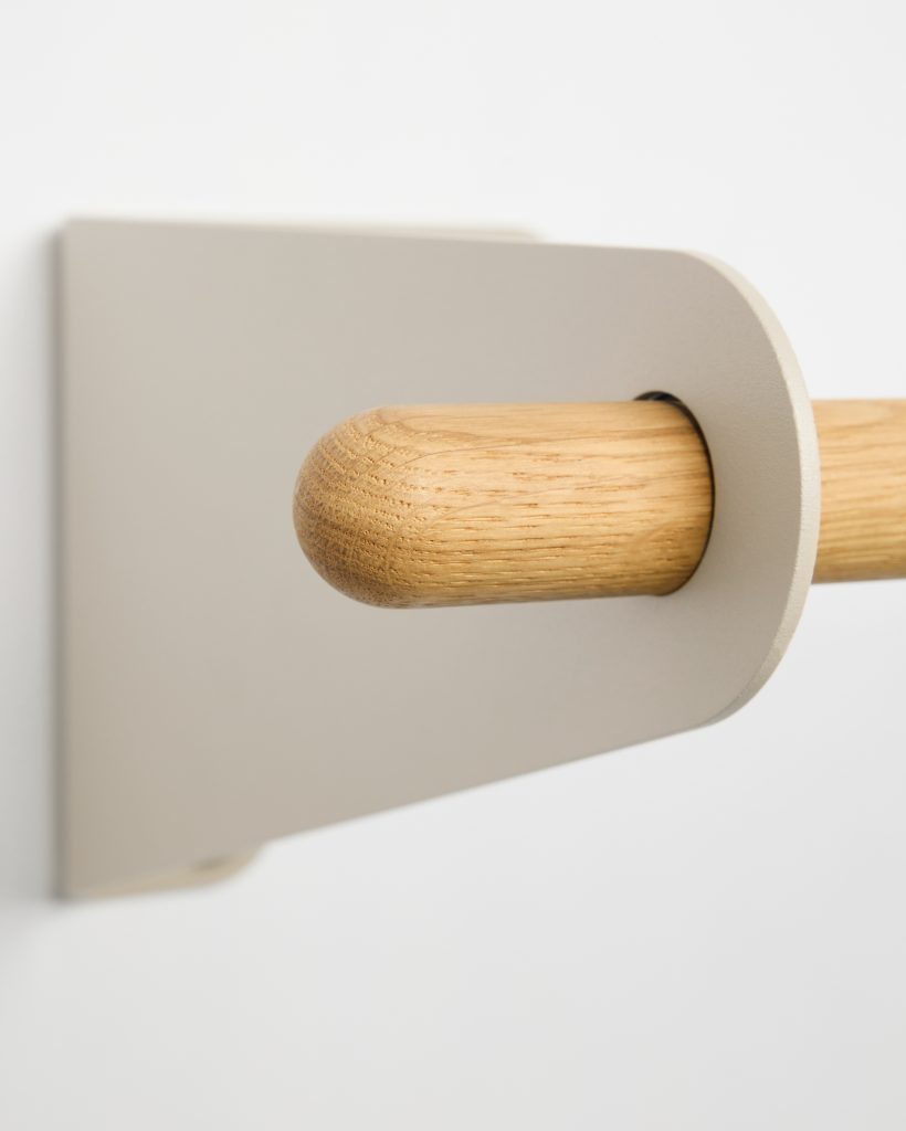 Close-up of a WoodUpp wall-mounted rail showing a natural oak dowel with rounded end fitted through a matte beige powder-coated metal bracket. The image highlights the wood’s fine grain and smooth finish and the minimalist rectangular wall plate with a rounded cut-out securing the dowel.
