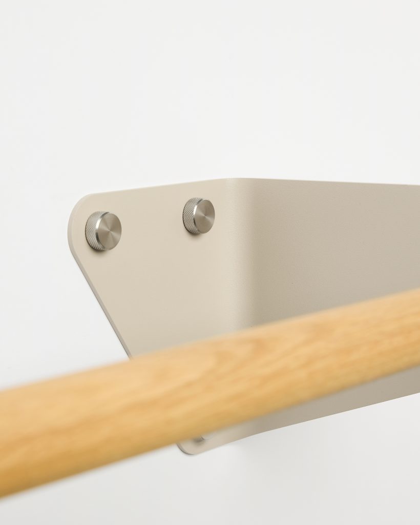 Close-up of WoodUpp wall bracket and oak barre: a WoodUpp powder-coated beige metal wall bracket fastened with two knurled stainless-steel screws, supporting a smooth, rounded light oak ballet barre with visible wood grain, shown mounted on a clean white wall in minimalist style.