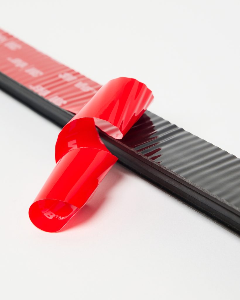 Close-up of a WoodUpp adhesive foam seal strip: a black, ribbed rubber foam strip with a glossy red peel-off protective backing partially removed to reveal sticky adhesive. The image highlights texture, layered foam edge and bright red release liner curling across a white background.