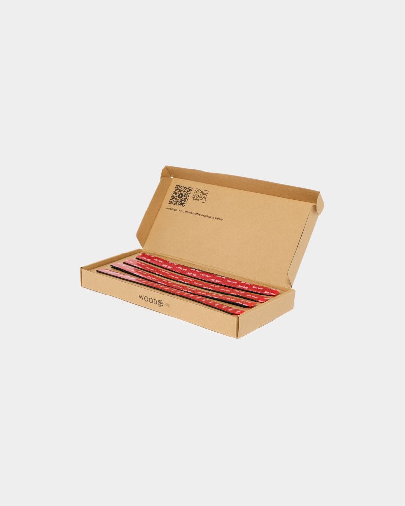 Open WoodUpp cardboard box showing four slim black Pop-On profile mounting strips with red 3M adhesive backing neatly laid inside. The kraft packaging bears WoodUpp branding and a QR/link to the Pop-On profile installation video, presenting the adhesive-backed profiles ready for installation.
