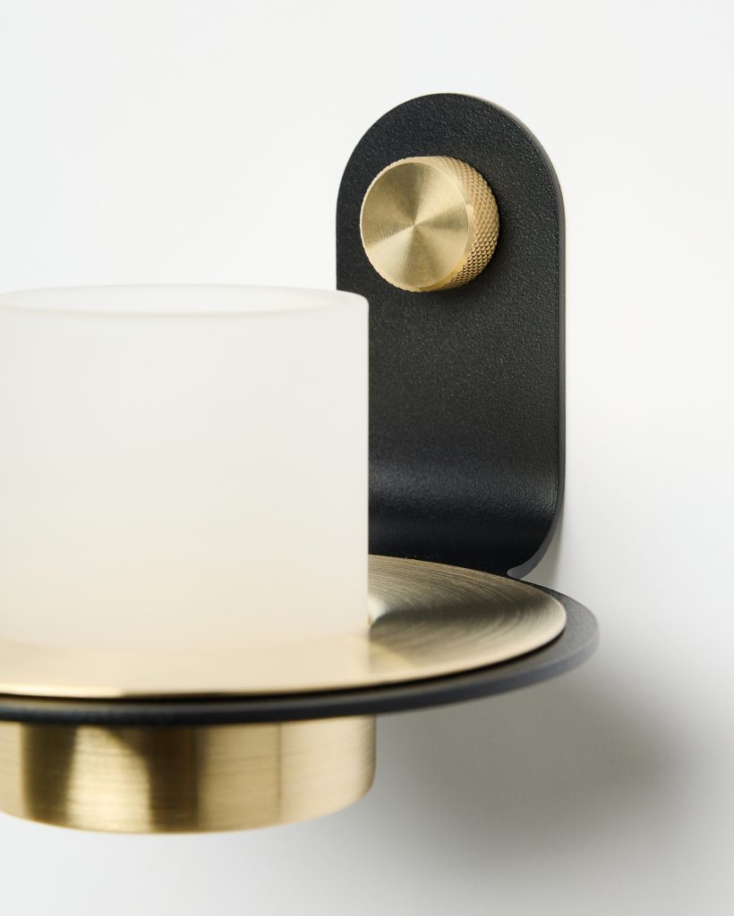 WoodUpp wall sconce featuring a matte black curved bracket, a knurled brass control knob, a brushed brass circular tray and cylinder base, and a frosted white cylindrical glass shade — a minimalist mixed-metal lamp highlighting WoodUpp's modern lighting design, quality craftsmanship.