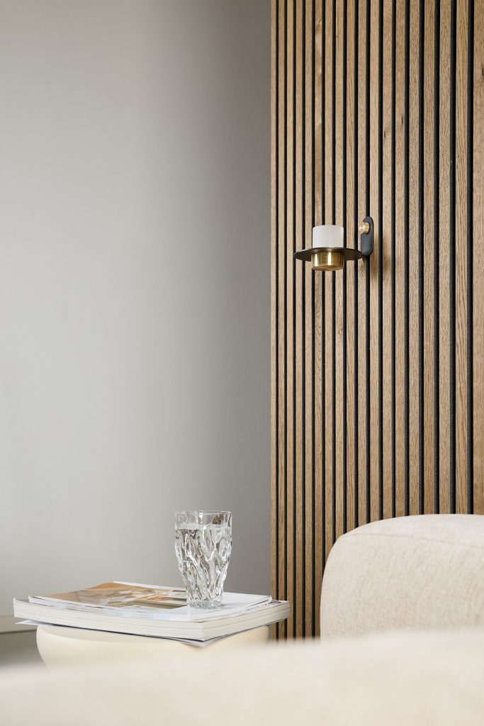 Minimal interior featuring WoodUpp vertical slatted oak wall panels on the right and a WoodUpp brass-and-black wall sconce holding a white frosted candle. In foreground, a beige upholstered sofa, a low round side table topped with stacked magazines and a textured clear drinking glass complete the scene.