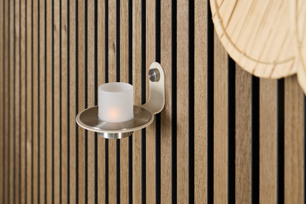 WoodUpp slatted oak wall panels fill the frame with vertical, evenly spaced light-oak slats and black acoustic gaps. A WoodUpp brushed-metal wall candle holder with a shallow dish and frosted glass LED votive is mounted to the panels; a woven bamboo wall decor peeks into the top-right corner.