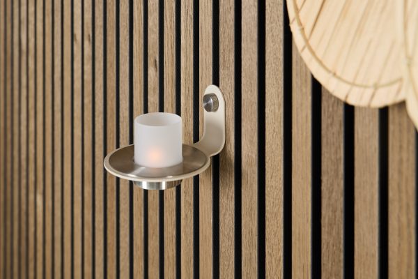 WoodUpp slatted oak wall panels fill the frame with vertical, evenly spaced light-oak slats and black acoustic gaps. A WoodUpp brushed-metal wall candle holder with a shallow dish and frosted glass LED votive is mounted to the panels; a woven bamboo wall decor peeks into the top-right corner.