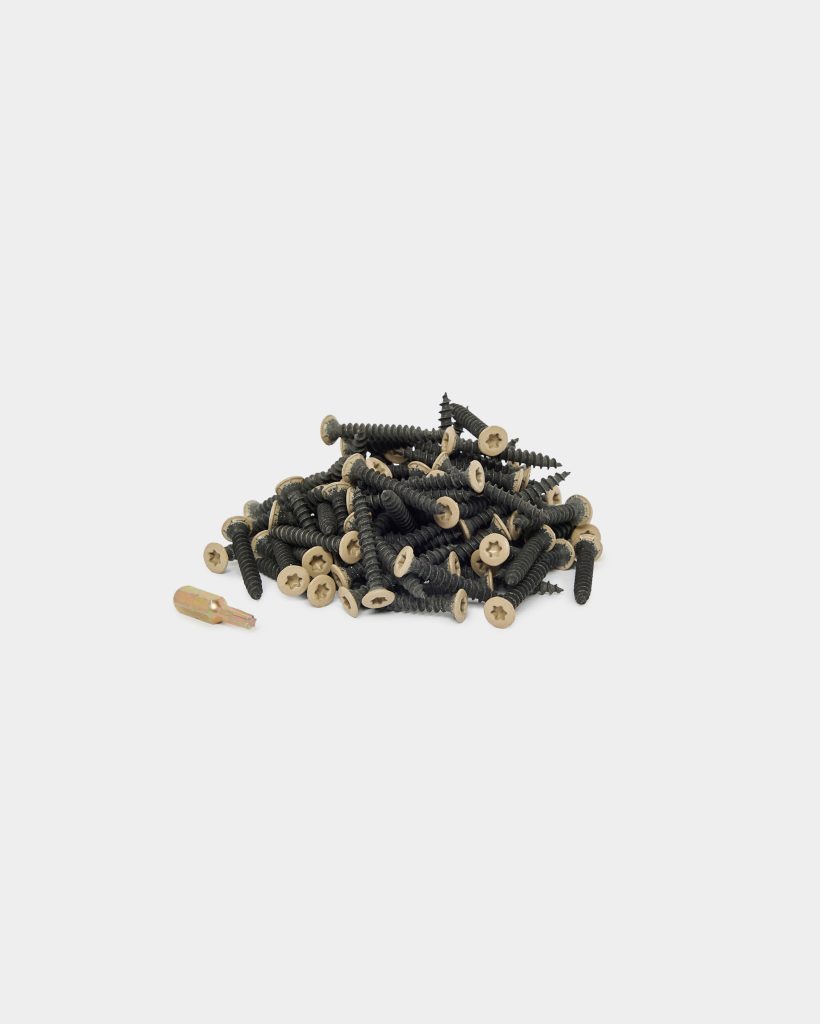 WoodUpp: pile of black coated wood screws with beige, countersunk star-drive heads and coarse threads, arranged in a low heap on a white background, accompanied by a single gold-colored driver bit. Products shown: WoodUpp decking/wood screws and matching impact driver bit.