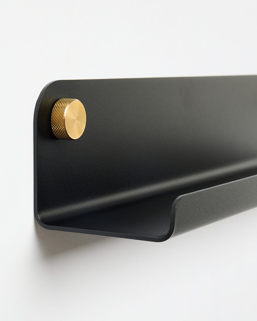 WoodUpp black wall shelf, a slim matte metal shelf with a curved front lip, shown mounted on a white wall using a round knurled brass fixing knob. The minimalist WoodUpp shelf and brass mounting detail emphasize clean lines and tactile metal finishes.