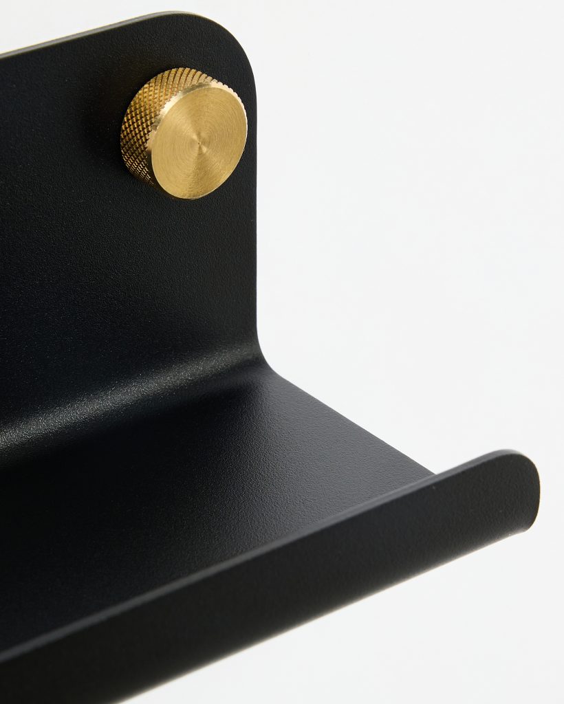 Close-up of a WoodUpp black powder-coated metal wall shelf bracket showing its angled shelf lip and rounded edge, secured with a knurled brass fixing knob. The textured matte surface and polished golden screw detail highlight WoodUpp’s minimalist, industrial storage accessory.