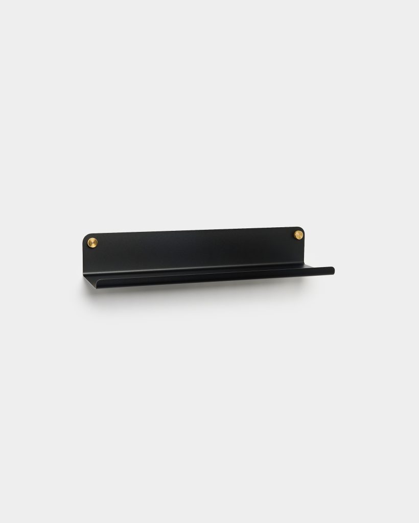 WoodUpp black wall shelf: a slim matte black metal floating shelf with a curved front lip and flat back plate, mounted with two round brass screws. Minimalist horizontal design suitable for small decor or toiletries, showing clean lines and concealed wall fixings.