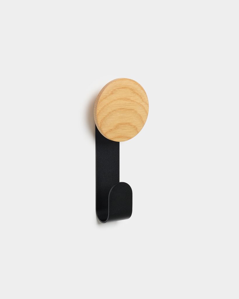 WoodUpp wall hook featuring a circular light-oak wooden knob with visible grain mounted atop a matte black metal hook that curves into a rounded lower crook, shown fixed on a plain white wall. Minimal, modern coat or bag hook from WoodUpp for hallway storage.