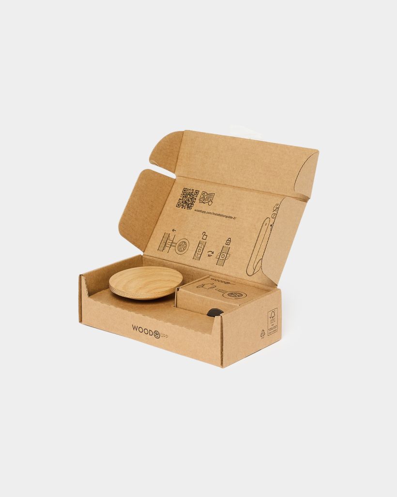 Open WoodUpp cardboard box with WoodUpp logo, displaying printed assembly icons and QR code, containing a round light-oak wooden coaster or shallow dish and a small boxed accessory. The WoodUpp-branded recyclable packaging neatly presents the natural-finish wooden product and a compact compartmented insert for parts.