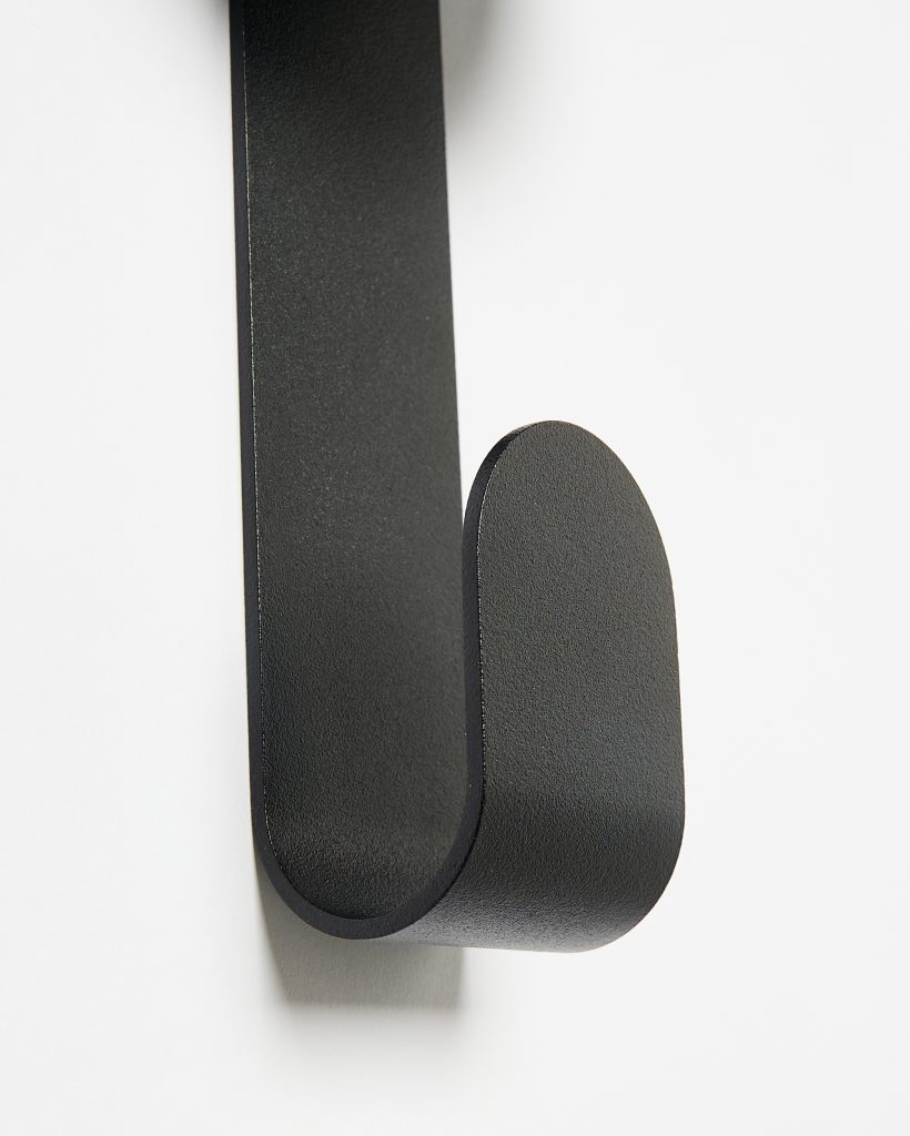 Close-up of a WoodUpp black matte metal wall hook, showing its single curved loop and rounded tip. The minimalist, textured hook is mounted on a white wall, with a slim vertical backplate and smooth, sturdy profile designed for hanging coats, towels or bags.