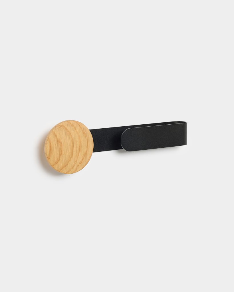 WoodUpp wall hook featuring a round oak disc peg mounted on a slim matte-black metal strap loop, shown fixed to a white wall; minimalist Scandinavian-style design with visible wood grain and smooth painted metal finish, casting a soft shadow to the left.