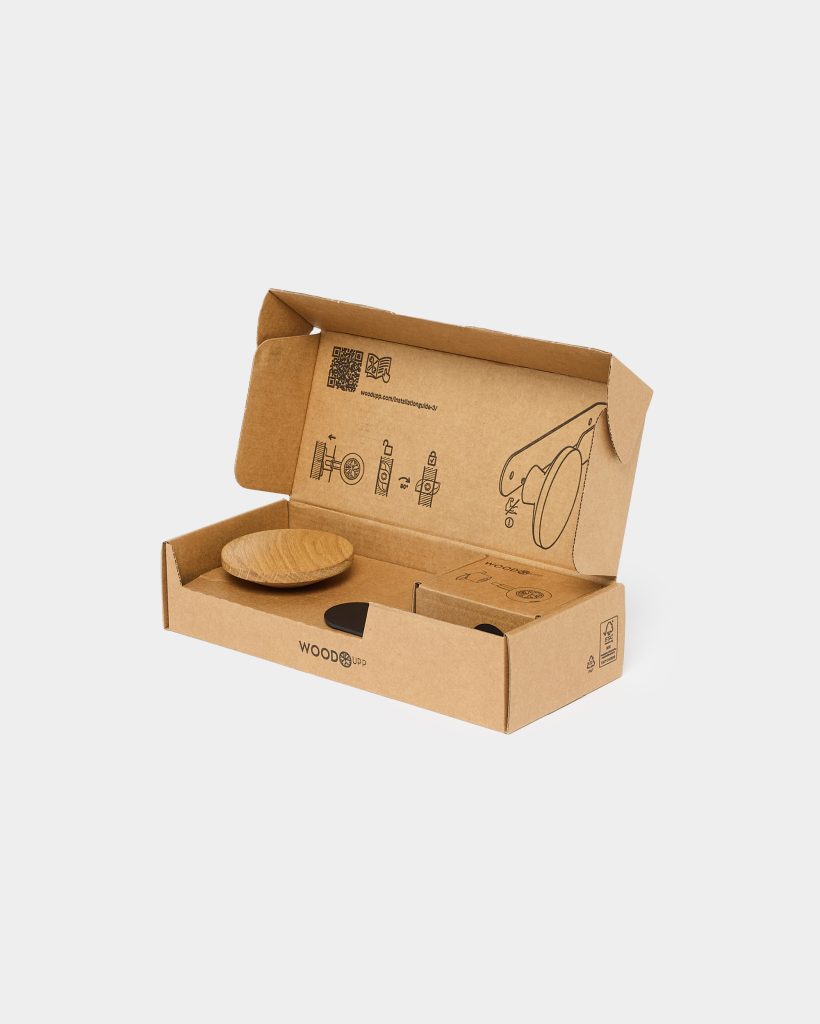 Open WoodUpp cardboard box showing a round oak wooden hook disc, a black adhesive mounting pad and a small hardware box nestled inside. Printed installation diagrams appear on the inner lid. Sustainable kraft packaging bears the WoodUpp logo and eco symbols.
