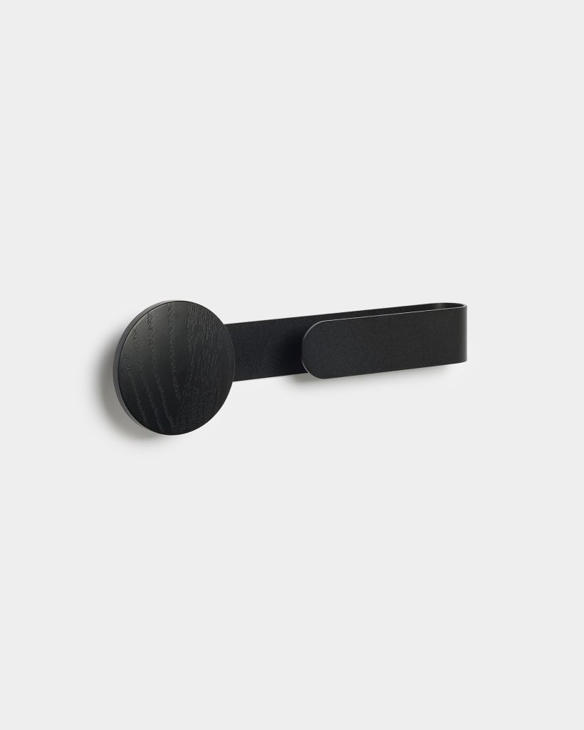 WoodUpp black wall hook featuring a circular black-stained wooden peg mounted to the wall, paired with a slim matte-black metal strap that curves into a practical hook. Minimalist, modern design shows wood grain texture on the round disc and a flat elongated metal loop for hanging.