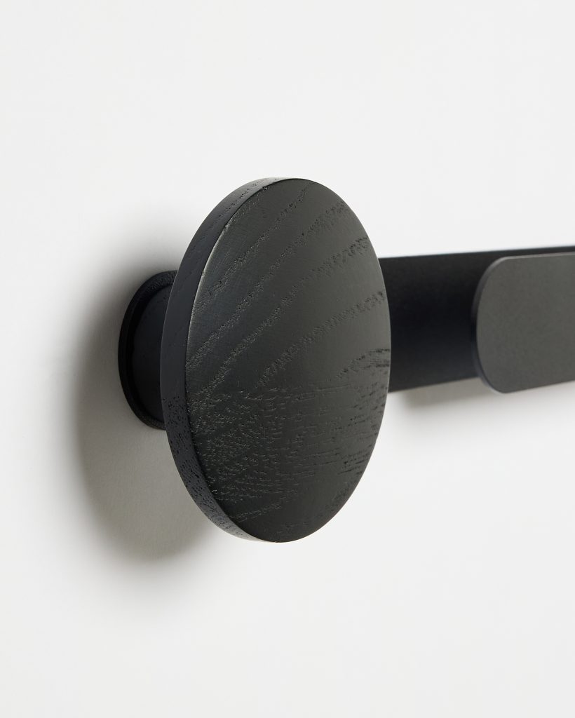 Close-up of WoodUpp wall-mounted coat rail showing a matte black wooden round peg hook and matching rectangular peg on a slim black metal rail. The wooden disc displays visible wood grain and textured finish, casting a soft shadow on the white wall behind.