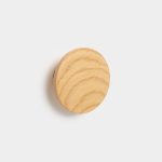 WoodUpp round wooden wall hook in light oak, shown mounted on a white wall. The circular, smooth-faced hook displays clear oak grain and a beveled edge, projecting a soft shadow. Minimal, natural finish highlights the wood texture — a simple, modern coat or accessory peg from WoodUpp.
