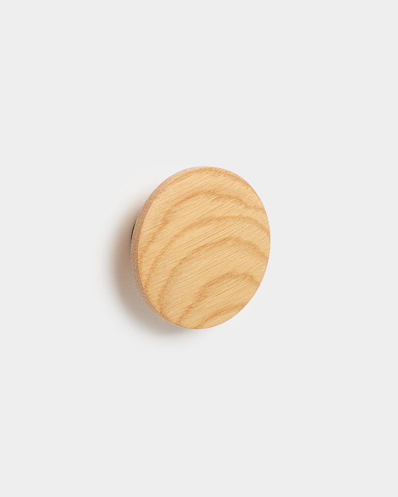 WoodUpp round wooden wall hook in light oak, shown mounted on a white wall. The circular, smooth-faced hook displays clear oak grain and a beveled edge, projecting a soft shadow. Minimal, natural finish highlights the wood texture — a simple, modern coat or accessory peg from WoodUpp.