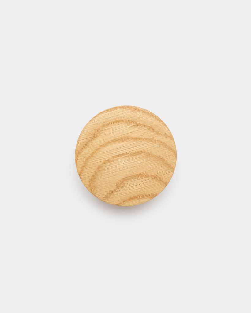 WoodUpp round wooden wall hook: a single circular light-oak wooden knob with visible natural grain and smooth sanded surface, centered on a clean white background and casting a faint shadow. The product is unadorned, showing concentric growth rings and warm honey tones, photographed from above to emphasize its round shape.
