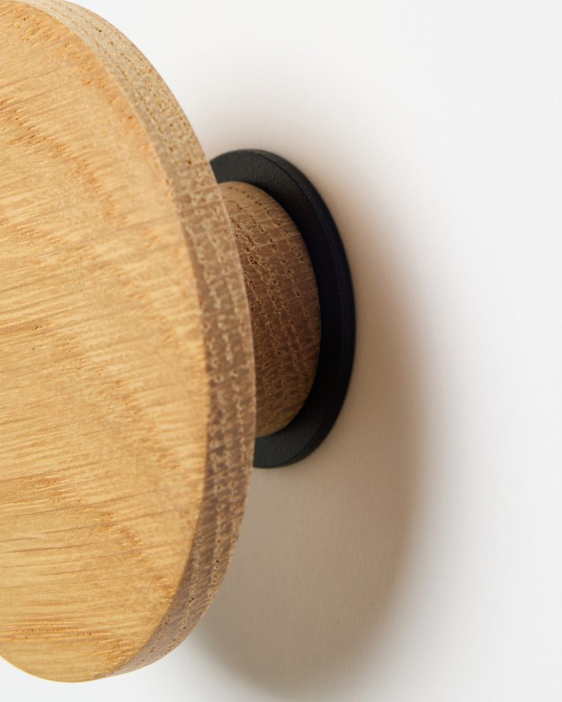Close-up of a WoodUpp wall hook: a round oak wooden peg with visible grain and layered edge, mounted on a matte black metal circular backplate against a white wall. The simple, natural wooden knob highlights handcrafted texture and minimalist Scandinavian design.