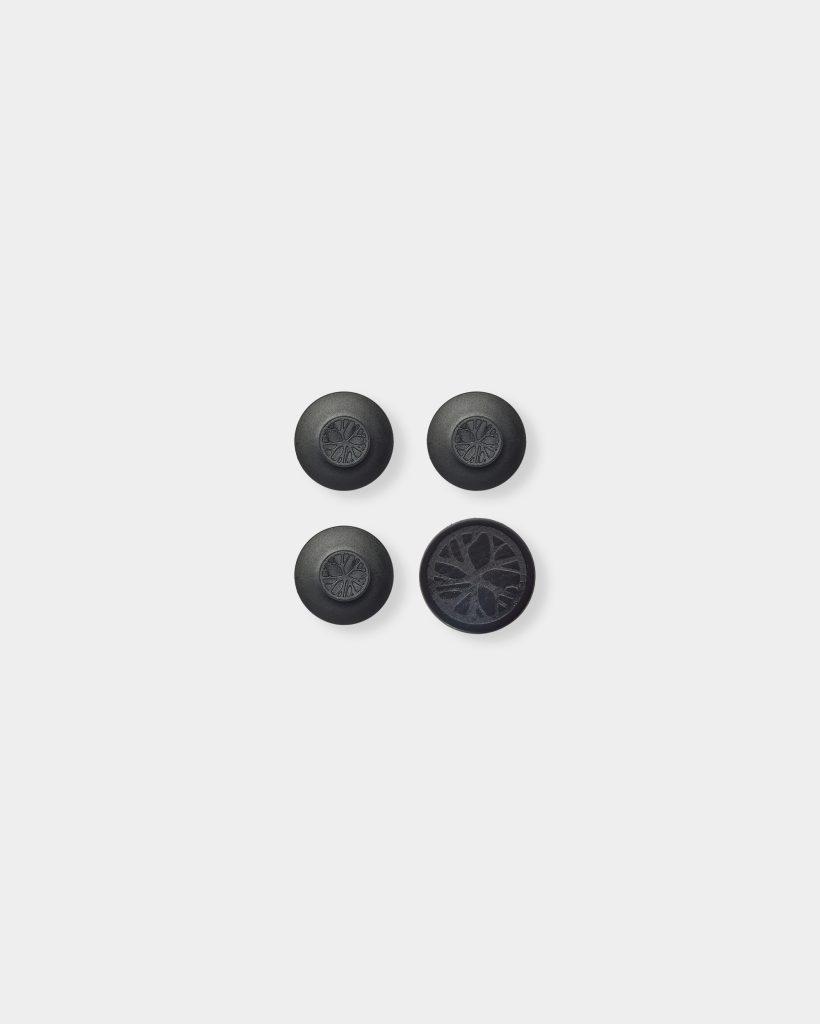 WoodUpp black rubber furniture bumpers shown on a white background: four round adhesive anti-slip pads with a subtle tree‑ring emblem. Three are slightly domed, one is a flatter disc revealing textured adhesive surface. Designed to protect surfaces, reduce noise and prevent sliding.
