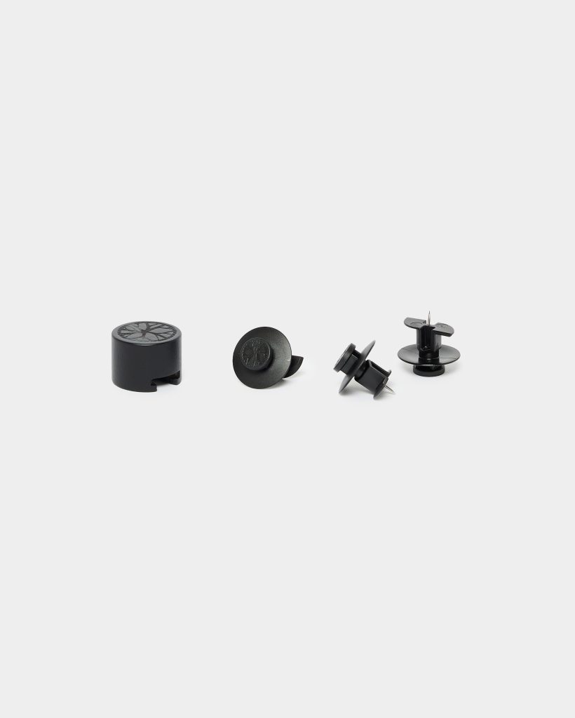 WoodUpp product photo showing four black plastic hardware pieces: a cylindrical cable-cap grommet with side slot, a round snap-in trim plug, and two push rivets/panel fasteners with metal pins and wide flanges, arranged in a neat row on a white background.
